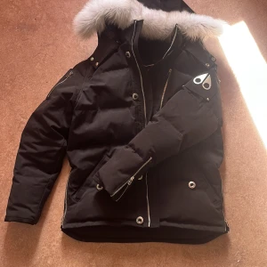 Svart dunjacka med pälskantad huva från Moose Knuckles - Säljer en svart dunjacka från Moose Knuckles med silverfärgade detaljer och pälskantad huva. Jackan har dragkedja, knappar och två fickor framtill. Klassisk logga på ärmen och snygg passform. Bra kvalitet bra form 
