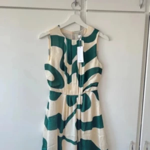 Grön och beige ärmlös klänning med abstrakt mönster - Säljer en snygg ärmlös klänning med grönt och beige abstrakt mönster. Klänningen har rund halsringning och markerad midja, perfekt för sommardagar. Lätt och luftig modell som ger en fräsch look. Prislapp på