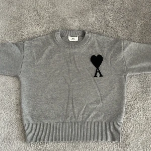 Grå stickad tröja från Ami - Säljer en grå stickad tröja från Ami med svart hjärta och bokstaven A på bröstet. Perfekt för en stilren och trendig look.