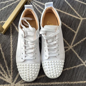 Vita sneakers med nitar från Christian Louboutin - Säljer ett par vita sneakers från Christian Louboutin med ikonisk röd sula och coola vita nitar på tån. Skorna har snörning och är tillverkade i skinn. Perfekta för dig som vill sticka ut med en lyxig och edgy stil.
