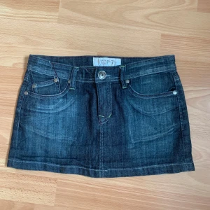 Mörkblå jeanskjol från Top 7 - Kort mörkblå jeanskjol med klassiska fickor fram och bak. Snygga broderade detaljer på bakfickorna och knappstängning framtill. Perfekt för en avslappnad och trendig stil. Längd 26 💗 midja tvärs över 35 💗