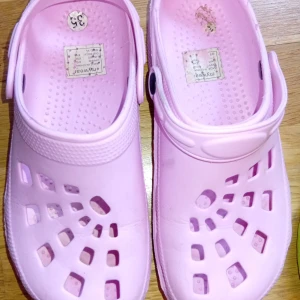 Ljusrosa tofflor i syntet - Säljer ett par ljusrosa tofflor i syntetmaterial med klassisk crocs-design. Skorna har ventilerande hål på ovansidan och justerbar hälrem. Perfekta för sommaren eller som inneskor.