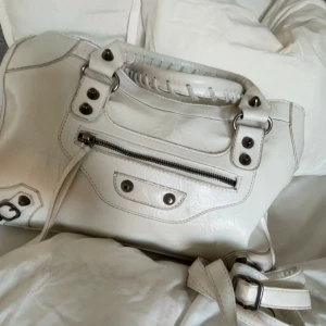 Vit handväska  - Säljer en snygg vit handväska som är köpt på plick, och vet då inte vilket märke den är ifrån. Liknar iallafall balenciagas citybag! Med silverfärgade metalldetaljer och dragkedja framtill. Väskan har både handtag och axelrem, samt dekorativa nitar och sömmar. Perfekt accessoar för att lyfta din outfit. ❣️