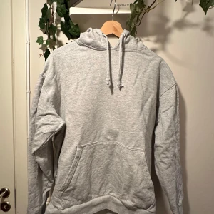 Ljusgrå hoodie från 157 - En enkel och stilren ljusgrå hoodie från 157 med magficka och justerbar huva med dragsko. Perfekt för en avslappnad look och passar till det mesta. Mjuk insida och ribbade muddar vid ärmslut och nederkant.