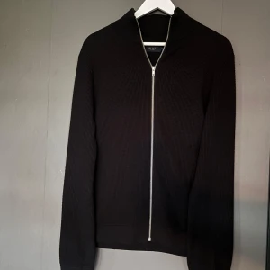 Svart stickad ziptröja från Riley - Säljer en svart stickad tröja från Riley med hel dragkedja framtill. Tröjan har lång ärm och en stilren, ribbad struktur. Perfekt för lager-på-lager och enkel att matcha med olika outfits.