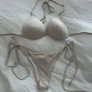 Beige ribbad bikini med knyt - Säljer en stilren beige bikini med ribbad struktur. Överdelen har vadderade kupor och knyt i nacken, medan underdelen har höga ben och knyt i sidorna. Perfekt för stranden eller poolen! Inte använd! 