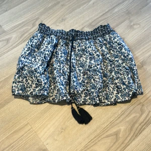 Blommig kortkjol från Zara - Supersöt kortkjol från Zara med blått och vitt blommönster. Kjolen har smock i midjan och knytband med tofsar framtill. Perfekt för en somrig look och lätt att matcha med olika toppar. De har även inbyggda shorts