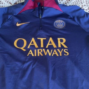 Paris Saint-Germain långärmad fotbollströja - Mörkblå långärmad PSG-tröja från Nike med gul logga och Qatar Airways-tryck på bröstet. Tröjan har dragkedja vid halsen och röda detaljer vid kragen. Perfekt för fotbollsfans som vill visa sitt stöd för Paris Saint-Germain.