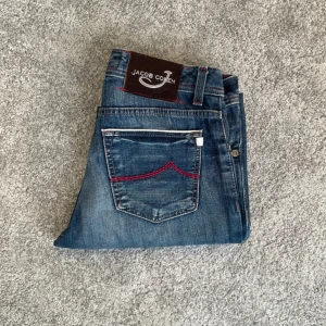 Jacob Choen Jeans Limited edition 688 (261 av 500) - Snygga blå jeans från Jacob Cohen med röda sömmar och detaljer på fickorna. Klassisk femficksmodell med knappar i gylfen och patch med logga bak. Jeansen har en rak passform och är tillverkade i denim. 100% bomull textil lim i anklen på byxorna syns inte utifrån 