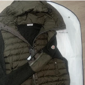 Olivgrön cardigan från Moncler med stickade ärmar - Säljer en olivgrön dunväst från Moncler med huva och stickade svarta ärmar. Västen har dragkedja framtill och Moncler-logga på ärmen. Perfekt för lager-på-lager och snygg detalj med blandat material.