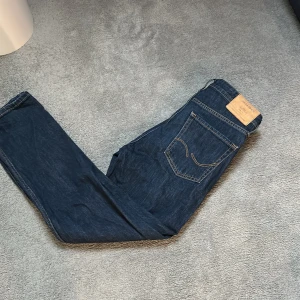 Mörkblå jeans från Jack & Jones - Säljer ett par mörkblå jeans från Jack & Jones med klassisk femficksdesign och läderpatch bak i midjan. Jeansen har raka ben och normal passform, perfekta till vardags. Snygga kontrastsömmar och robust denimkänsla.