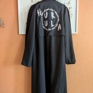 Trenchcoat med Hurula tryck från Monki  - Svart lång kappa från Monki med knappar framtill och unikt Hurula tryck på ryggen. Klassisk krage och rak passform, perfekt för dig som vill sticka ut med detaljer.skärp saknas den är baggy/boxig ish använd för en plåtning mycket bra skick