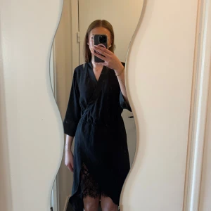 Svart omlottklänning med spets från Vila - Säljer en svart omlottklänning från Vila i storlek M. Klänningen har trekvartsärm, knyt i midjan och vacker spetsdetalj nedtill. Perfekt för dig som gillar stilrena och feminina plagg.