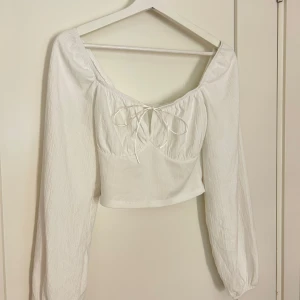 Vit croppad blus med knyt - Säljer en söt vit croppad blus med långa ärmar och knyt framtill. Blusen har en lätt rynkad struktur och en djupare urringning. Perfekt att styla med jeans eller kjol för en trendig look.
