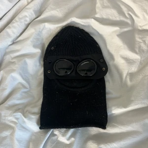 CP COMPANY BALACLAVA - Säljer en svart stickad balaclava med inbyggda glasögon från C.P. Company. Balaclavan är helsvart och har en ribbad struktur. Perfekt för dig som vill ha en unik och stilren accessoar.