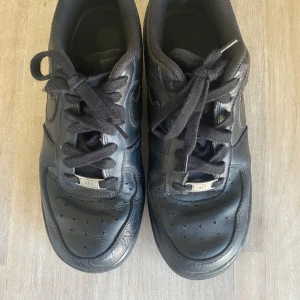 Svarta Nike Air Force 1 sneakers - Klassiska helsvarta Nike Air Force 1 sneakers med svart snörning och rund tå.  Skorna är knappast använda då de är för stora! Perfekta för vardagen, fina och väldigt sköna!