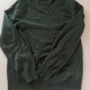 Mörkgrön v-ringad tröja från J.Lindeberg - Mörkgrön v-ringad tröja från J.Lindeberg med diskret logga på bröstet. Tröjan har långa ärmar och ribbade muddar vid ärmslut och nederkant. Perfekt för lager-på-lager under kyligare dagar. Liten defekt till höger på magen. Se bild