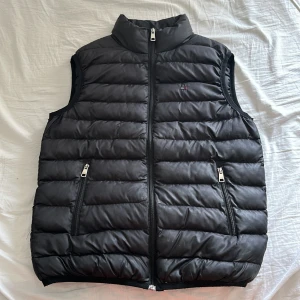 Svart dunväst från Tommy Hilfiger - Snygg svart dunväst från Tommy Hilfiger med quiltad design och dragkedja framtill. Västen har två fickor med dragkedja och en liten broderad logga på bröstet. Perfekt för lager-på-lager-stil.
