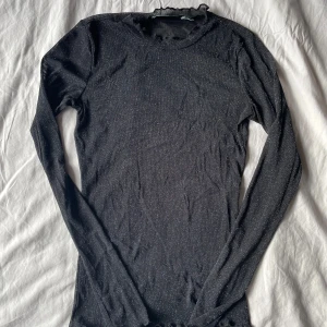 Svart glittrig mesh topp - Säljer en svart långärmad topp med glittrigt tyg och volangdetaljer vid ärmslut och nederkant. Tunt transparent och stretchigt material som ger en snygg passform. Perfekt att styla till olika outfits!