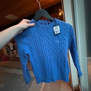 Blå kabelstickad tröja från GANT - Säljer en blå kabelstickad tröja från GANT med rund halsringning och diskret logga på bröstet. Tröjan har lång ärm och ribbade muddar vid ärmslut och nederkant. Perfekt för en stilren och klassisk look.