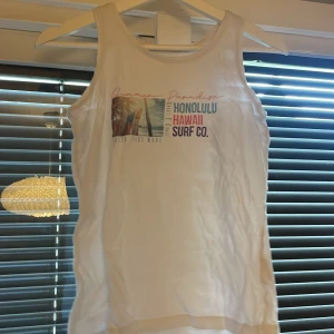 Vit linnetopp med tryck från Reaction - Säljer en vit linnetopp från Reaction med färgglatt tryck framtill som har texten 'Honolulu Hawaii Surf Co.' och strandmotiv. Perfekt för sommaren och strandhäng.