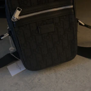 Svart axelväska från Christian Dior - Snygg svart axelväska från Christian Dior med justerbar rem och silverfärgad dragkedja. Väskan har ett diskret mönster med Dior-logga över hela ytan och en praktisk ytterficka. Perfekt för att bära det viktigaste med stil.