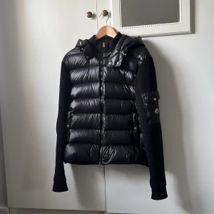 Moncler puffer jacka - Säljer en svart pufferjacka från Moncler med huva och stickade ärmar. Jackan har dragkedja framtill, quiltad framsida och en ficka med Moncler-logga på ärmen. Perfekt för kyliga dagar och snygg till många outfits.