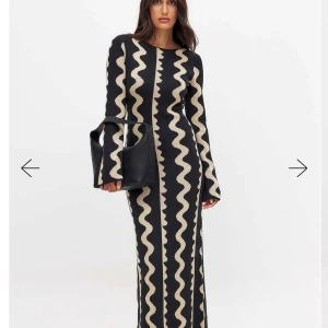 Jacquardstickad långklänning med vågigt mönster - Säljer en svart långklänning med beige vågigt jacquardmönster. Klänningen har lång ärm och figurnära passform. Tillverkad i stickat material som ger en snygg siluett. Helt oanvänd med lappen kvar, nypris 799kr