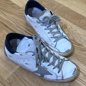 Vita sneakers från Golden Goose med grå stjärna - Säljer ett par vita sneakers från Golden Goose med grå mockadetaljer och en grå stjärna på sidan. Bra skick överlag dock tecken på användning på hälarna (se bilder) 