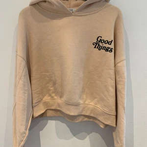 Beige hoodie med tryck 'Good Things' - Säljer en beige hoodie med svart texttryck 'Good Things' fram och bak. Tröjan har huva, långa ärmar och är croppad i modellen. Perfekt för en avslappnad stil.