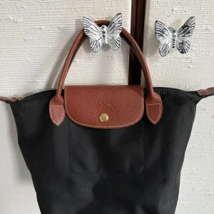 Svart handväska från Longchamp - Klassisk svart handväska från Longchamp med bruna skinnhandtag och lock. Väskan har guldfärgad knapp med logga och dragkedja upptill. Perfekt storlek för vardagsprylar och enkel att bära i handen eller på armen.