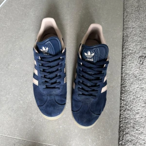 Adidas Gazelle blå sneakers - Säljer ett par klassiska Adidas Gazelle sneakers i blå mocka med vita ränder och beige detaljer. Skorna har platt sula, snörning och rund tå. Perfekta för en stilren och sportig look. Skorna har en slitning vid sulkanten på vänster sko men inget som syns när man använder dom. Storlek 46!