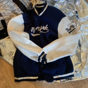 Blå och vit collegejacka med New York-tryck - Snygg blå och vit collegejacka med broderat New York-tryck på bröstet och ärmen. Jackan har knappar framtill, randiga muddar och klassisk varsity-stil. Perfekt för en sportig och trendig look.