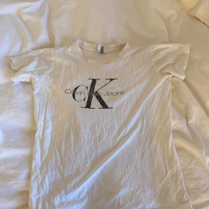 Vit t-shirt med Calvin Klein Jeans tryck - Vit t-shirt med stort svart Calvin Klein Jeans-tryck på bröstet. Klassisk rund hals och korta ärmar. Mjuk och tunn bomullskvalitet, perfekt för en enkel och stilren look.