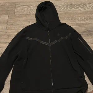 Svart huvudtröja Nike Teck  - Svart hoodiejacka från Nike med dragkedja framtill och reflekterande detaljer över bröstet och på ärmen. Jackan har huva och en ficka med dragkedja på ena ärmen. Perfekt för en sportig och stilren look. Nypris på dessa tröjor är 1300