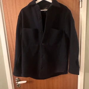 Svart overshirt med knappar - Säljer en stilren svart overshirt med knäppning framtill och två stora bröstfickor. Jackan har en klassisk krage och lång ärm, perfekt för lager-på-lager-stil. Passar dig som gillar enkel och tidlös design.