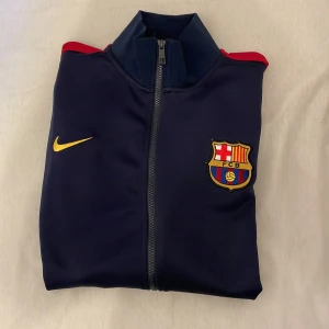 FC Barcelona träningsjacka från Nike - Mörkblå träningsjacka från Nike med röda detaljer och FC Barcelona-logga på bröstet. Gult Nike-märke och texten 'BARÇA' i rött på ryggen. Jackan har dragkedja och ståkrage, perfekt för fotbollsentusiasten.