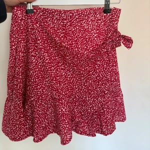 Röd kortkjol med vitprickigt mönster och volang - Superfin röd kjol med vita prickar och volang nertill. Kjolen har omlottkänsla med en söt rosett i sidan och är perfekt för sommarens alla tillfällen. Lätt och luftig modell som ger en skön känsla.