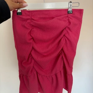 Rosa volangkjol med rynkade detaljer från Gina Tricot  - Säljer en snygg rosa kjol med volang och rynkade detaljer framtill. Kjolen har en kortare modell och är perfekt för dig som vill ha något trendigt och färgglatt. Materialet känns mjukt och har en lätt struktur. Kjolen har även en topp som jag säljer om man skulle önska det som ett sett. 