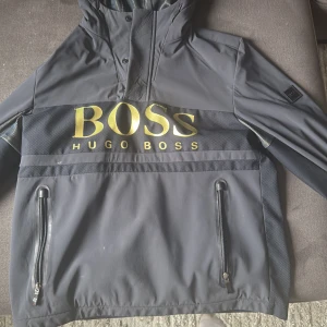 Svart vindjacka från Hugo Boss med huva - Säljer en svart vindjacka från Hugo Boss med stor guldfärgad logga. storlek mellan M-L