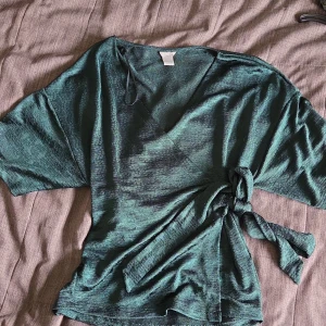 Grön omlottopp med knyt från Lindex - Snygg grön omlottopp från Lindex med knyt i midjan och korta ärmar. Toppen har en v-ringad hals och är tillverkad i ett mjukt, lätt material som ger en avslappnad känsla.