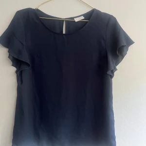 Mörkblå blus med volangärm från Vila - Säljer en stilren mörkblå blus från Vila med korta volangärmar. Blusen har rund halsringning och en liten knapp i nacken. Perfekt för dig som gillar en enkel men elegant look.