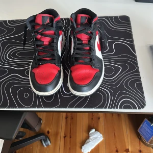 Nike Air Jordan 1 i rött, svart och vitt - Säljer ett par klassiska Nike Air Jordan 1 med högt skaft i färgerna rött, svart och vitt. Skorna har svart snörning, rund tå och platt sula. Perfekta för dig som gillar streetwear och sneakers med ikonisk design.