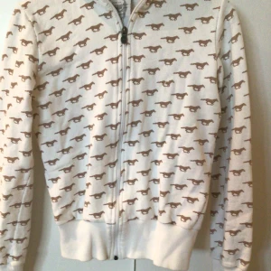 Vit hoodie med brunt hästmönster från Jenny Hellström - Vit hoodie med dragkedja och brunt hästmönster över hela tröjan. Mjukt material och ribbade muddar vid ärmslut och nederkant. Perfekt för dig som gillar unika prints och vill sticka ut.