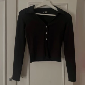 Svart ribbad kofta från Divided - Säljer en svart ribbad kofta från Divided i storlek XS. Tröjan har långa ärmar, krage, v-ring och knäppning framtill med tre knappar. Perfekt att styla till olika outfits. Aldrig använd men slet bort lappen