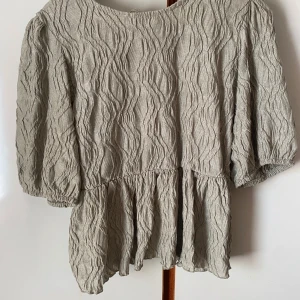 Grön topp med struktur och knyt i ryggen - Säljer en grön topp från gina tricot med vågig struktur, puffiga trekvartsärmar och snygg knytning i ryggen. Toppen har en lös passform och är perfekt för dig som gillar unika detaljer.