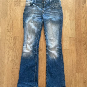 Blå bootcut jeans med slitna benslut - Säljer ett par blå bootcut jeans med slitna benslut och ljusa tvättade partier framtill och baktill. Jeansen har klassisk femficksdesign, normal passform och stängs med dragkedja och knapp.