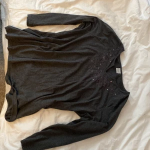 Mörkgrå långärmad topp med broderi från Florie - Säljer en mörkgrå långärmad topp från Florie i storlek XXL. Toppen har v-ringning och vackert broderade detaljer med små glittriga stenar framtill vid halsen. Perfekt för dig som gillar diskreta men fina detaljer.