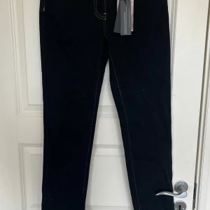 Mörkblåa jeansbyxor från Versace med guldiga detaljer - Mörkblåa jeansbyxor från Versace med guldiga sömmar och ikonisk Medusa-broderi på bakfickan. Byxorna har en guldig knapp framtill med Versace-logga och raka ben. Klassisk femficksmodell med exklusiv känsla.