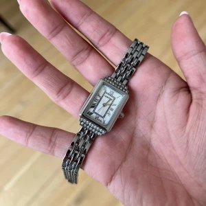 Silverfärgad klocka - Säljer en elegant rektangulär klocka i silverfärg med länkarmband. Urtavlan har romerska siffror och är dekorerad med gnistrande stenar runt boetten. Perfekt accessoar för att lyfta din stil.